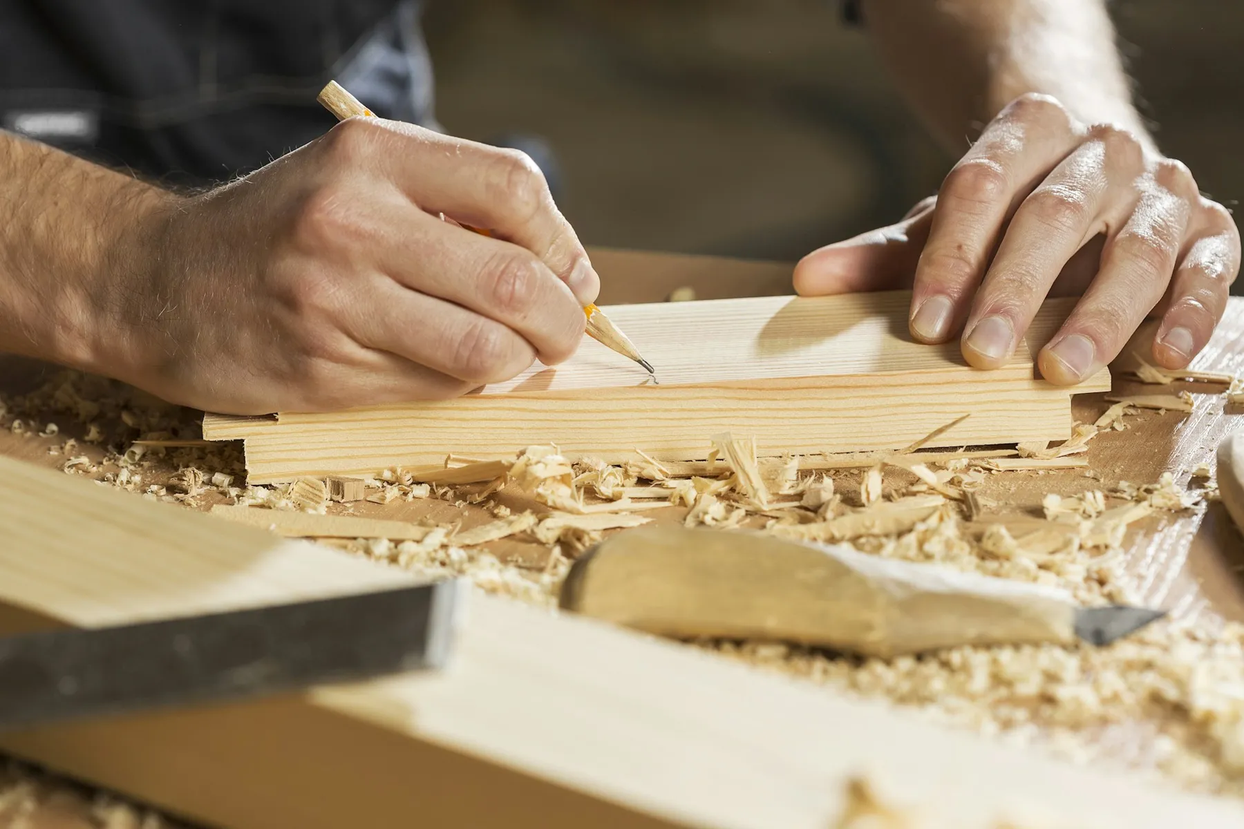 Photo d’ambiance : mains d’artisan traçant au crayon sur une planche de bois, copeaux sur l’établi